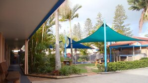 A covered outdoor area with a concrete floor, wooden benches, and a fenced garden with palm trees. - Yamba Twin Pines Motel (Yamba)