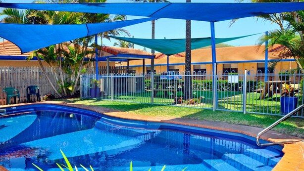 A swimming pool with a blue water feature, surrounded by a fenced area with palm trees and a building with a red roof in the background. - Yamba Twin Pines Motel (Yamba)