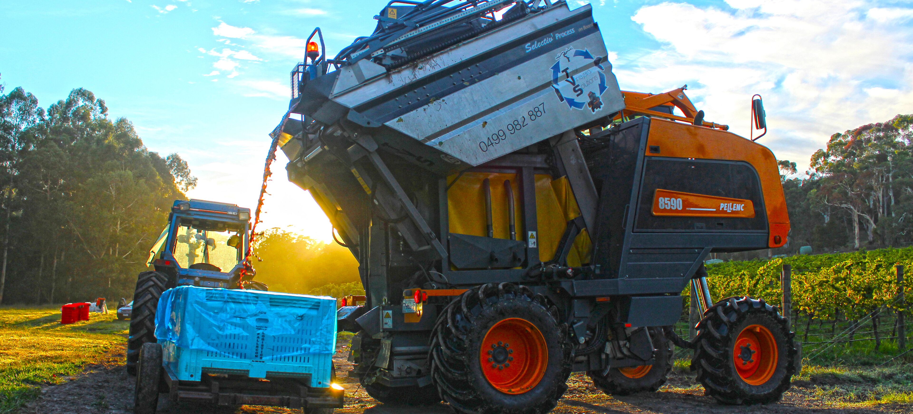 Une grande machine agricole, avec un conteneur bleu à l’avant, est garée sur un chemin de terre, avec des arbres en arrière-plan.