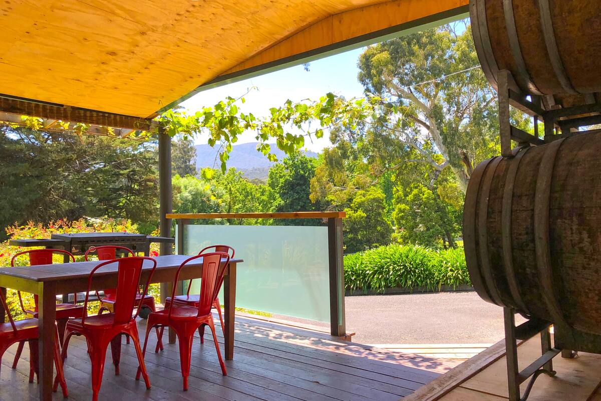 An outdoor patio with a wooden table and red chairs, a glass enclosure, and a view of greenery and mountains.
