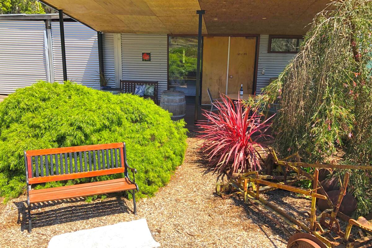 A covered outdoor seating area with a bench, a planter with red foliage, and a wheelbarrow.