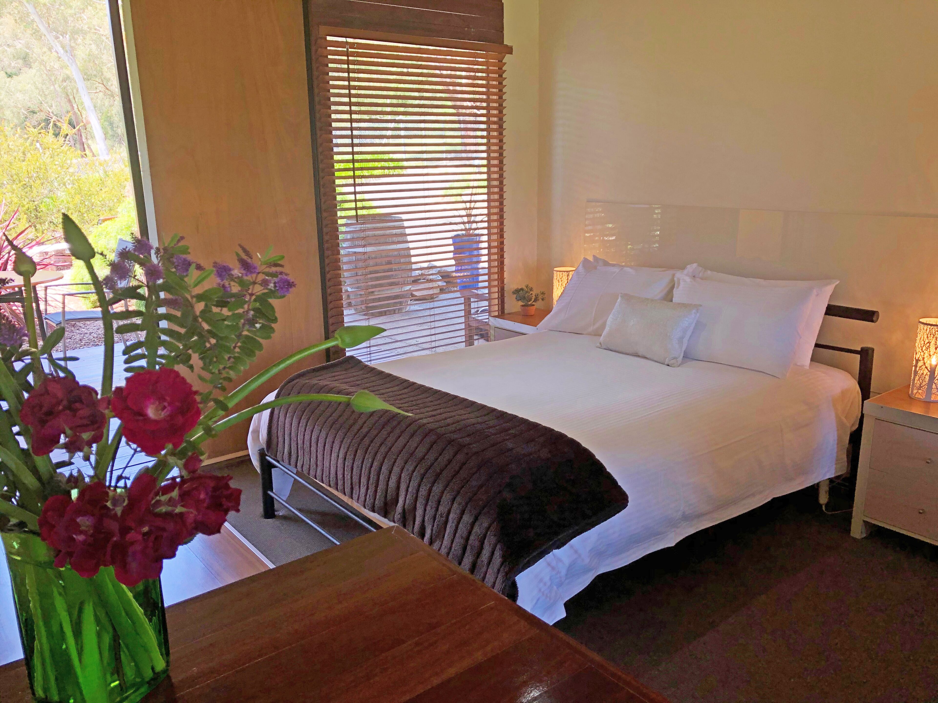 Une chambre à coucher comprenant un lit, des tables de chevet, une commode, une fenêtre avec des stores et un vase avec des fleurs.