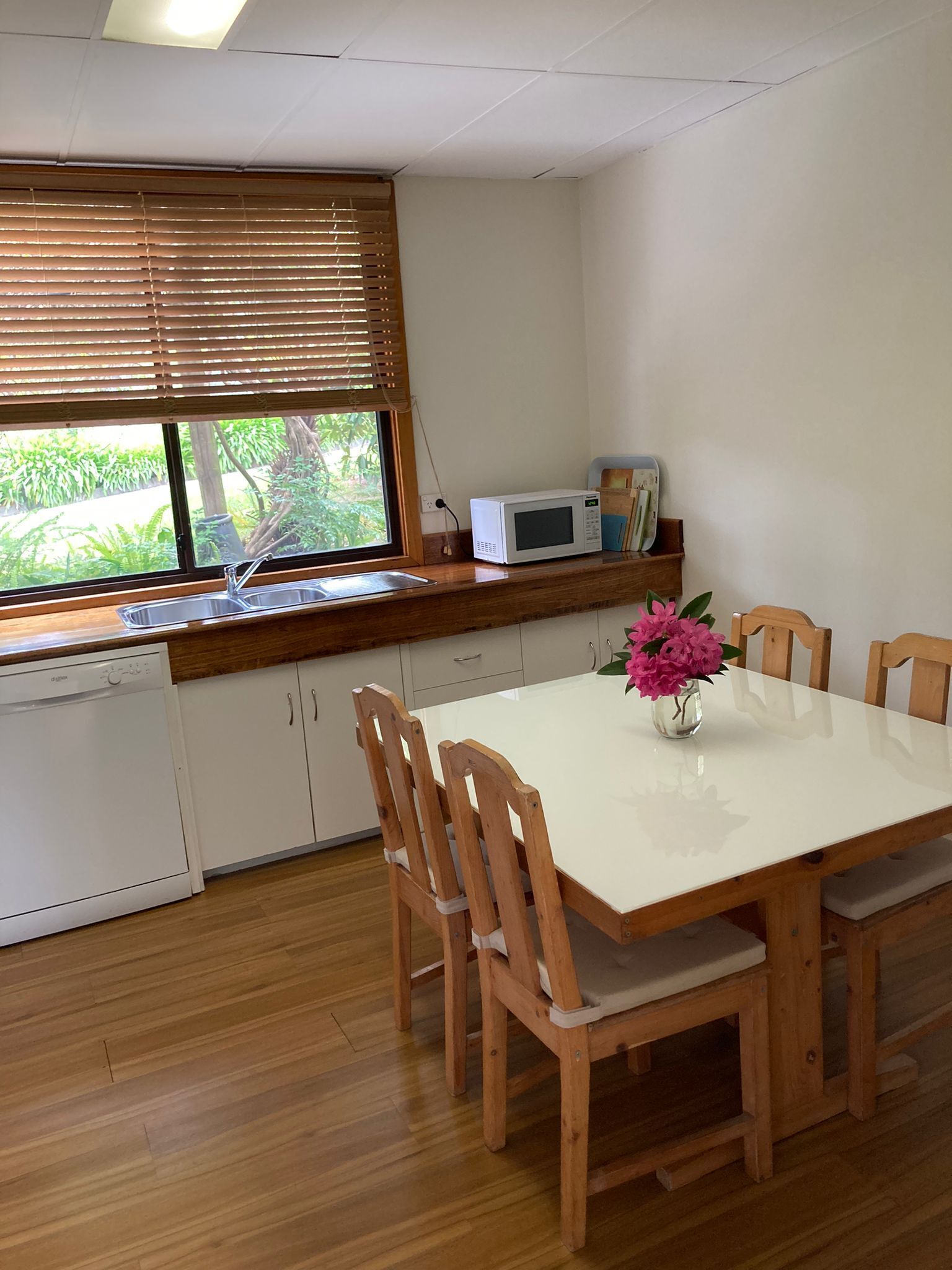 A kitchen with a white countertop, wooden cabinets, a white microwave, and a window with blinds.