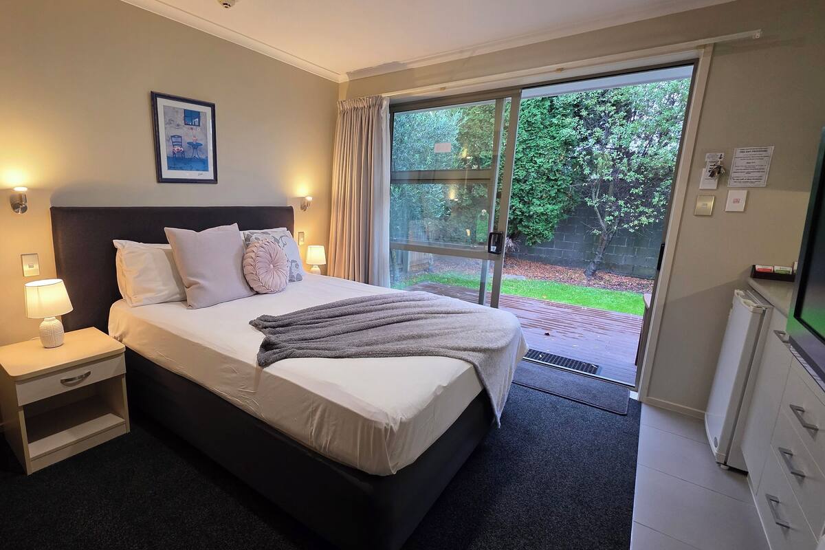 A hotel room with a bed, bedside tables, a lamp, a framed picture, and a view of the outdoors.