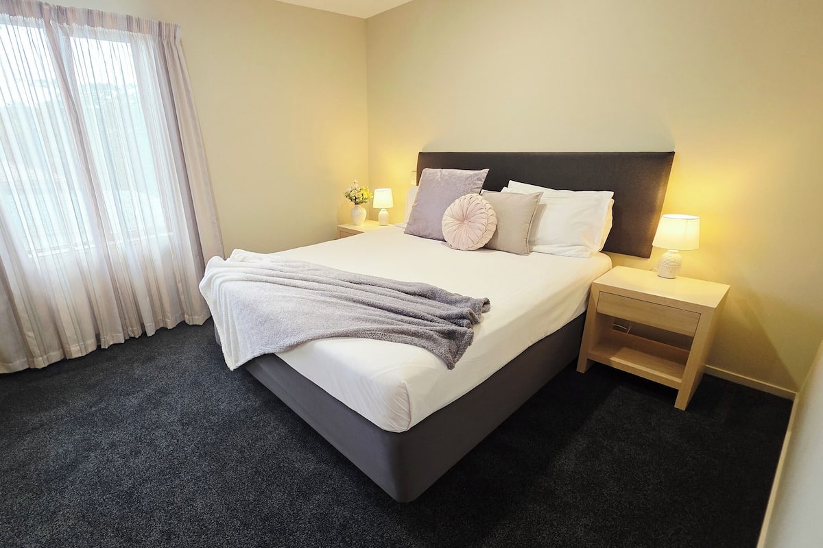 A hotel room with a large bed, two bedside tables with lamps, a dark carpet, and a window with sheer curtains.