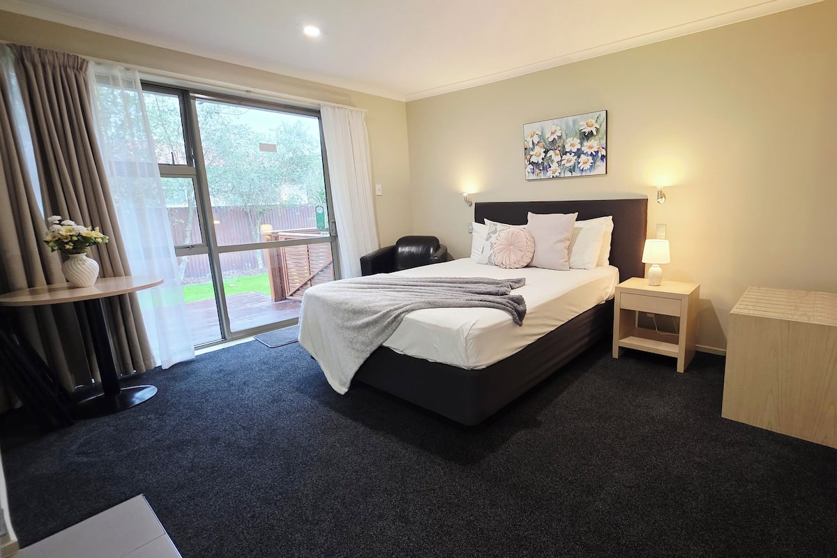 A hotel room with a large bed, a bedside table with a lamp, a small wooden cabinet, and a sliding glass door leading to an outdoor area.