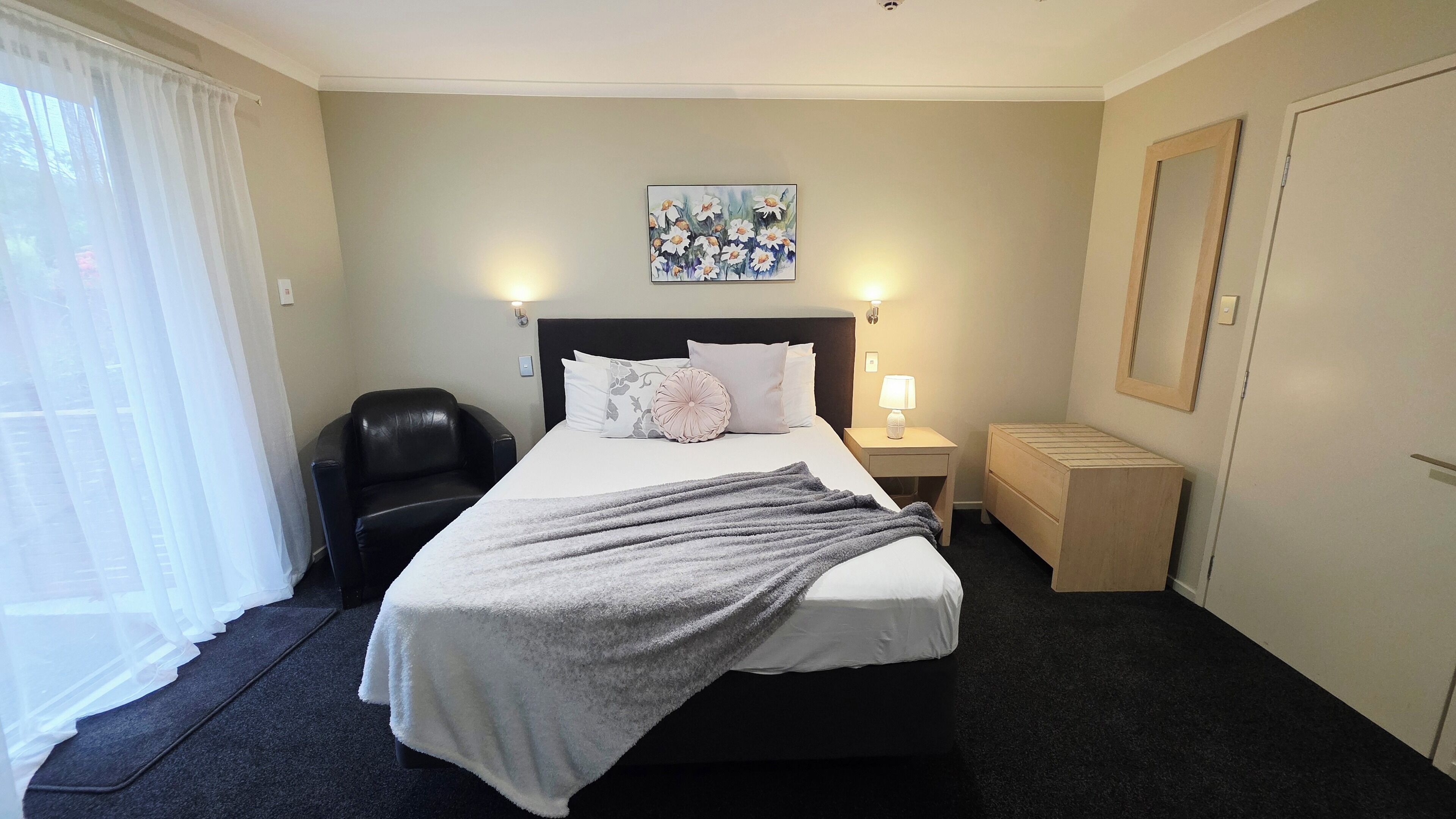 A hotel room with a bed, a chair, a nightstand, a lamp, a painting on the wall, and a window with curtains.