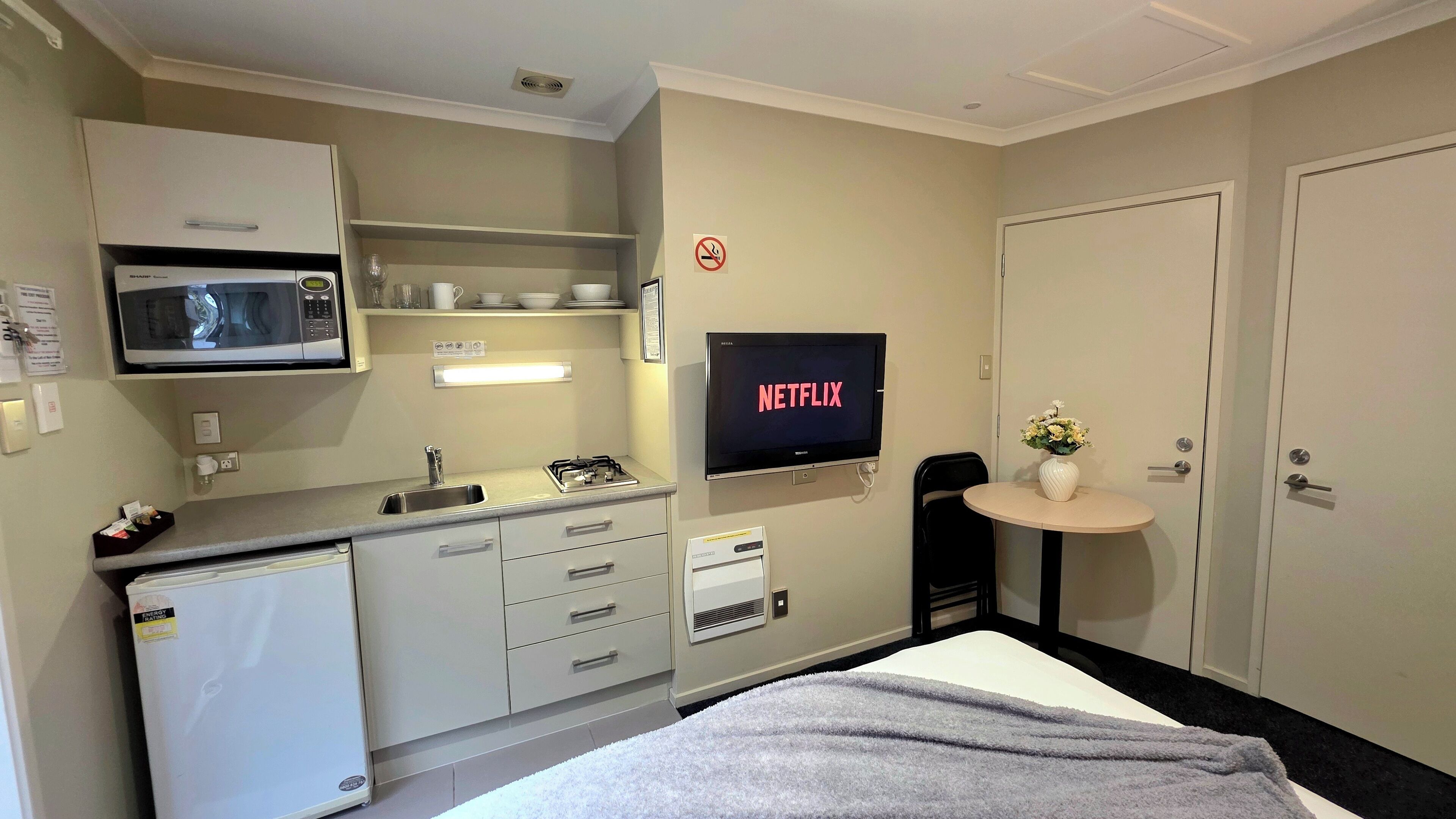 A hotel room with a kitchenette, a TV displaying 'NETFLIX', a no-smoking sign, a small table with a vase, and a bed with a grey blanket.