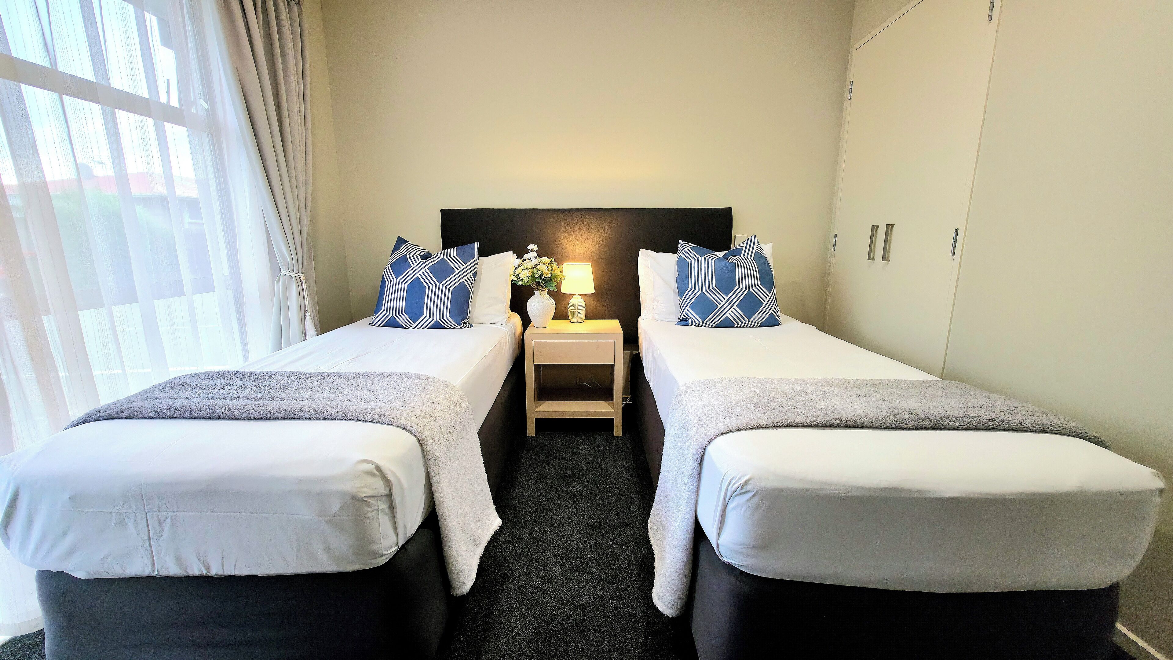 A hotel room with two single beds, a nightstand with a lamp, and a window with curtains.