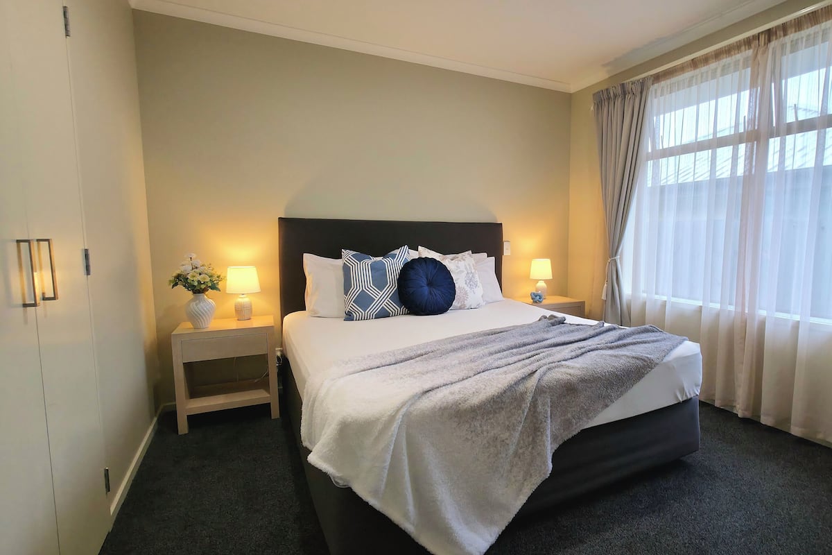 A hotel room with a large bed, two bedside tables with lamps, a nightstand, a window with sheer curtains, and a wall-mounted light fixture.