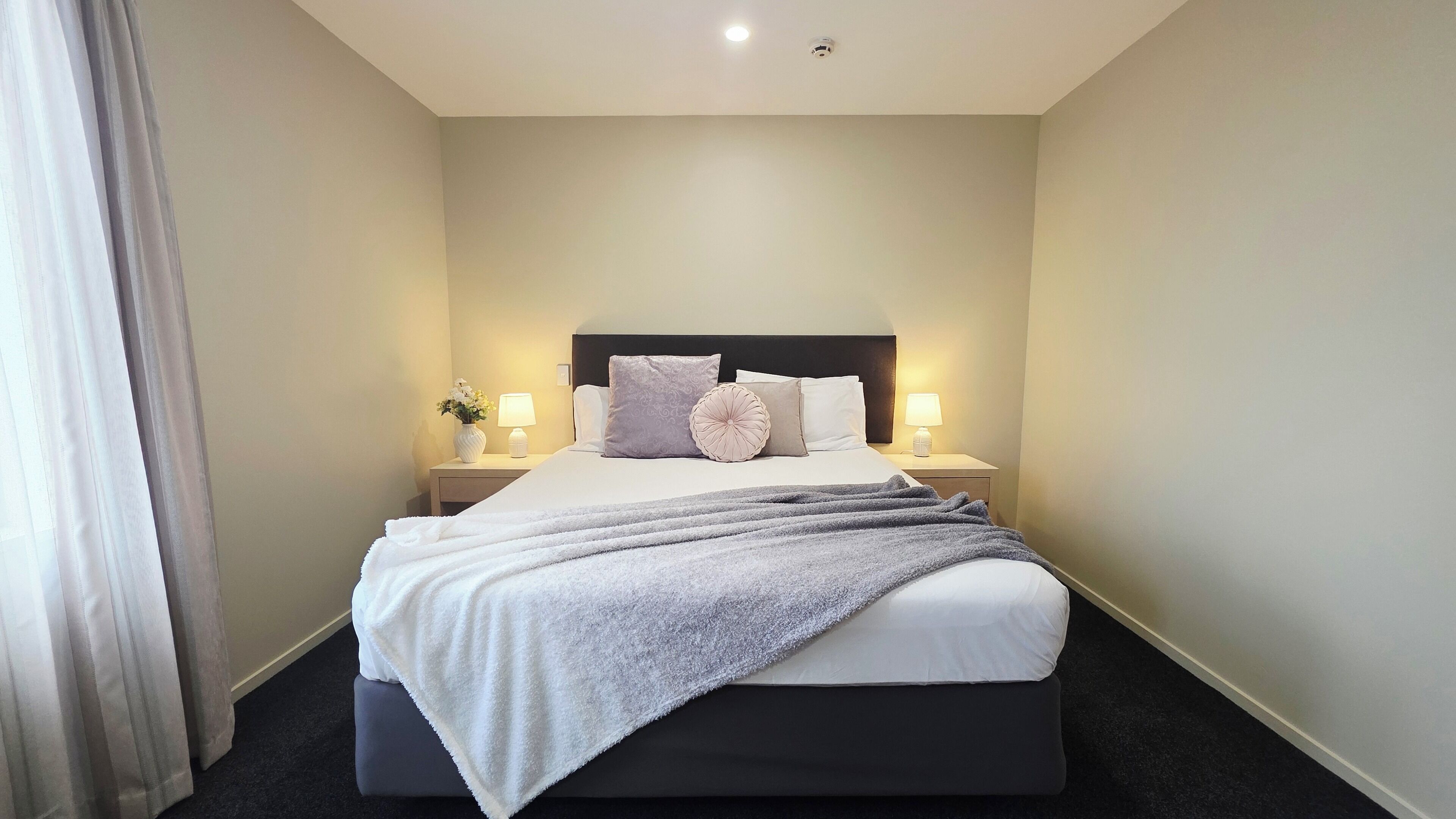 A neatly made bed with a grey blanket, two bedside lamps, and a vase with flowers on the nightstand.