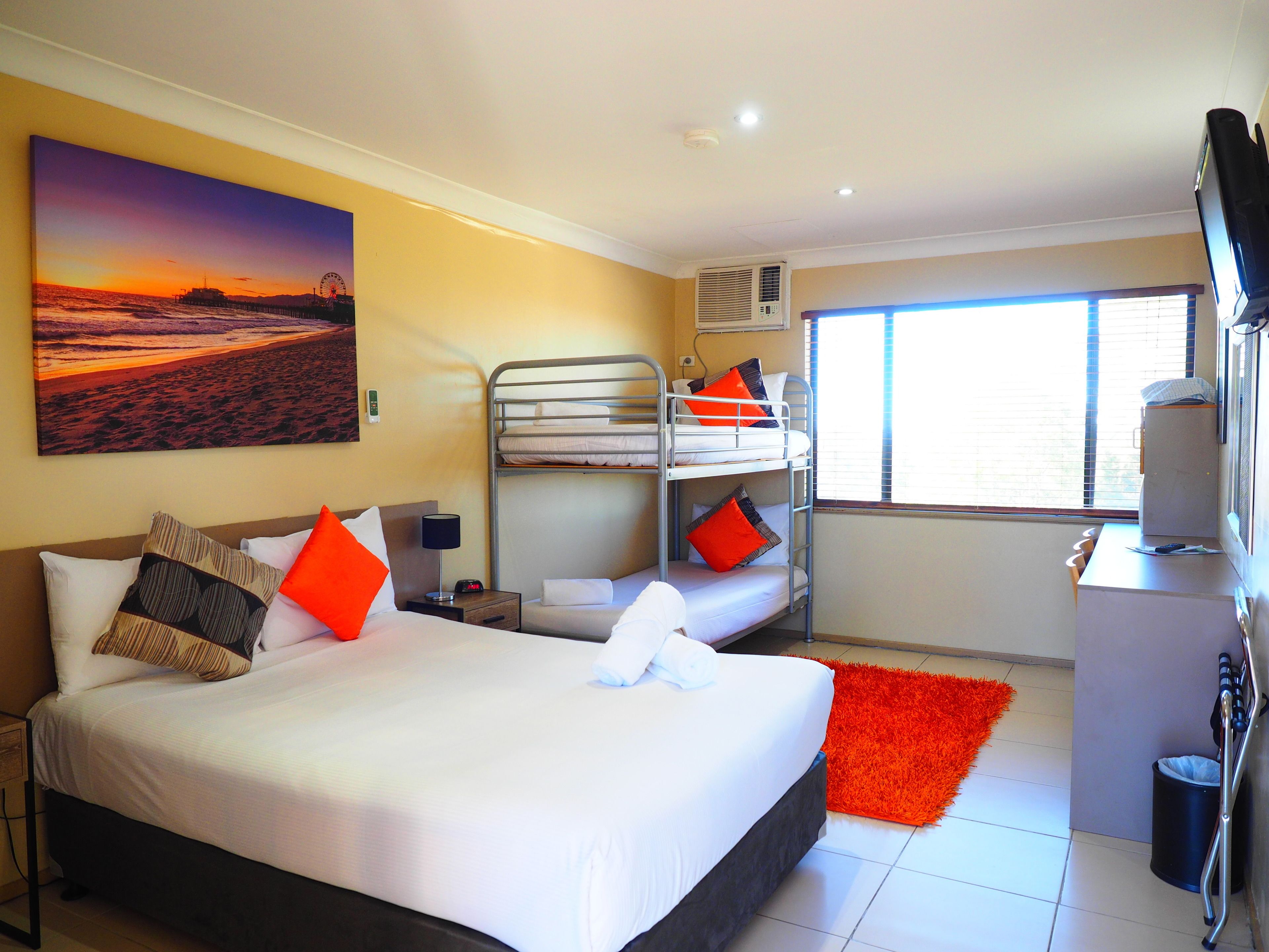 A hotel room with a large bed, a bunk bed, a desk with a computer, a TV, and a window with blinds.