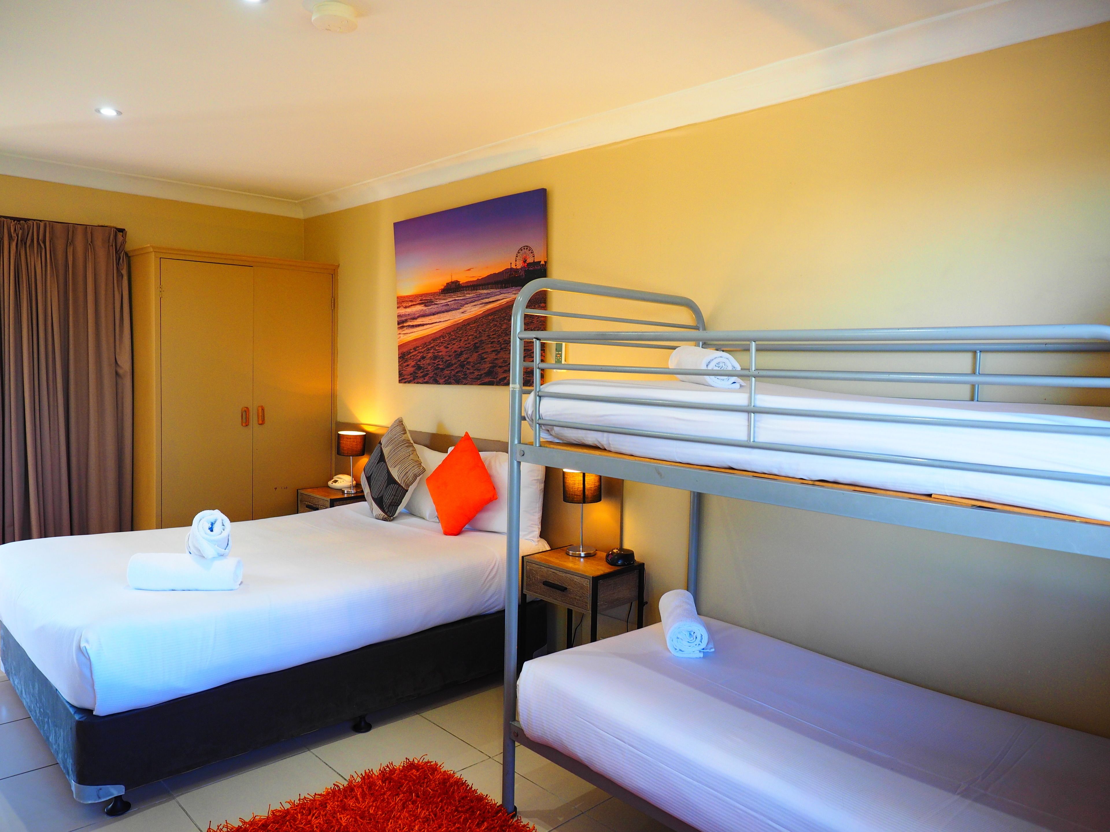 A hotel room with two beds, one with a bunk bed and the other with a single bed. There is a painting on the wall, a lamp on the side table, and a red rug on the floor.