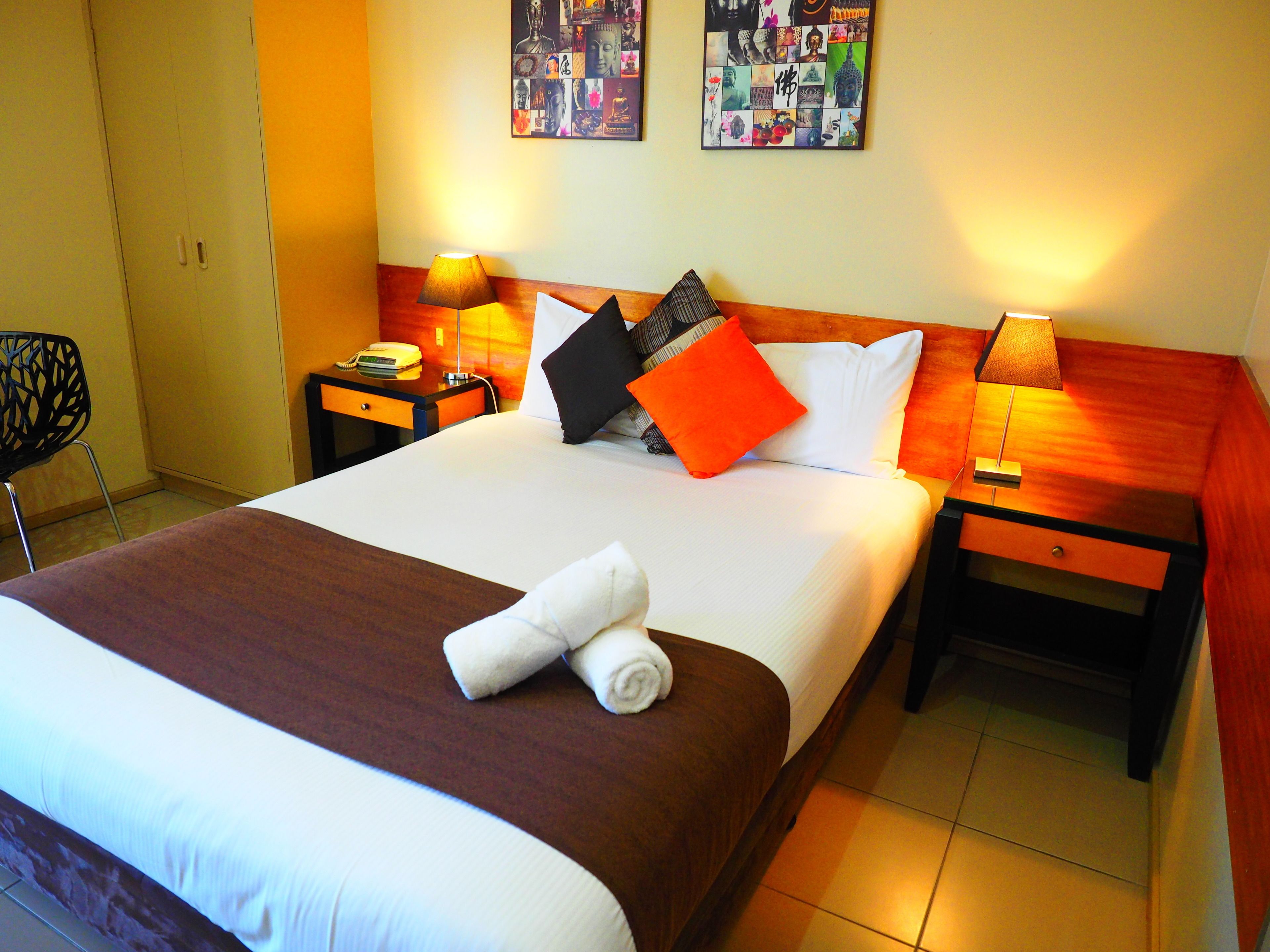 A neatly made bed with a brown bedspread, white pillows, and an orange accent pillow. There are two bedside tables with lamps, a chair, and a wall with artwork.