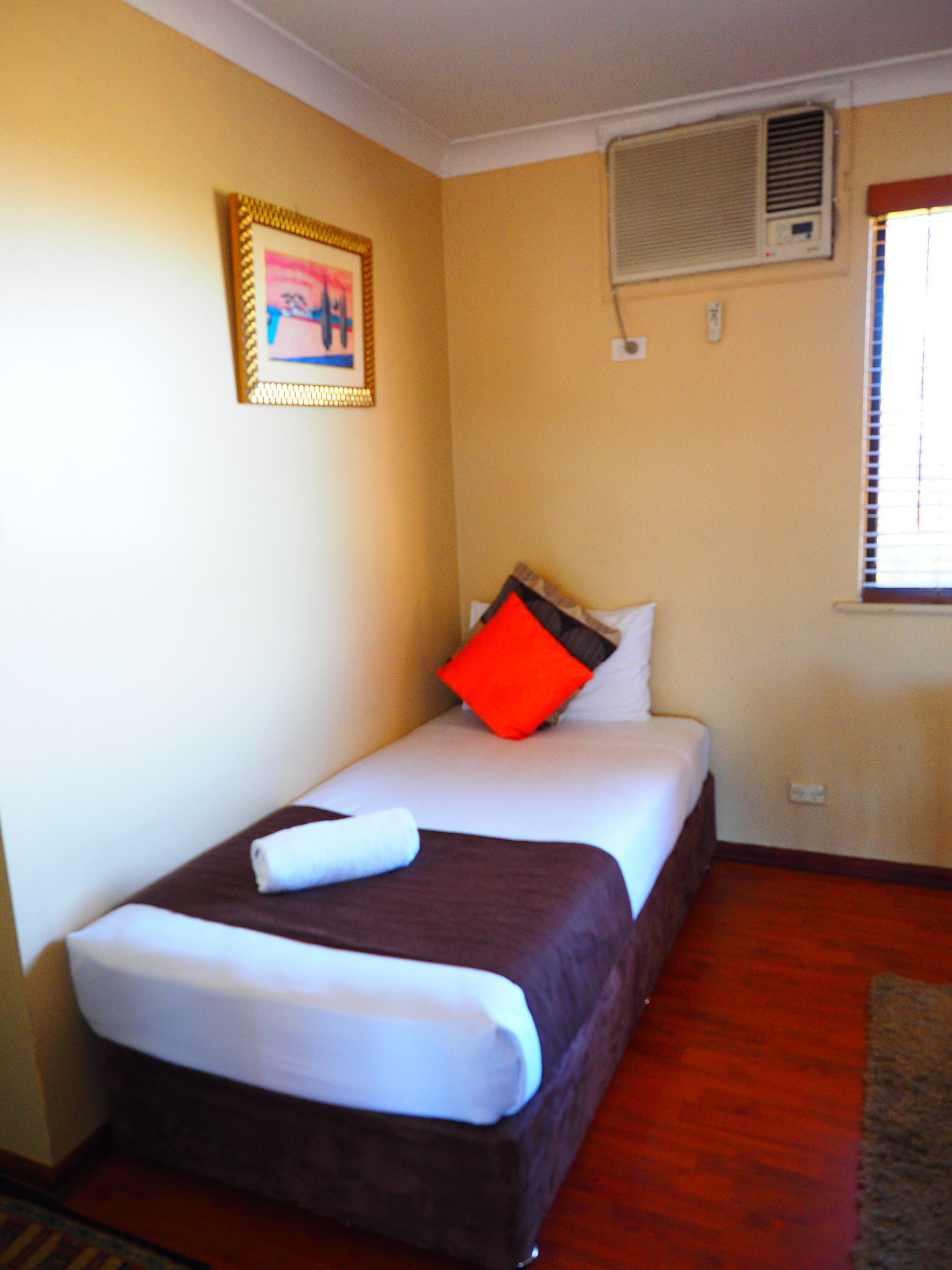 A small, neatly made bed with a brown headboard, white bedding, and a red pillow. There is a framed picture on the wall above the bed, and an air conditioning unit mounted on the wall.