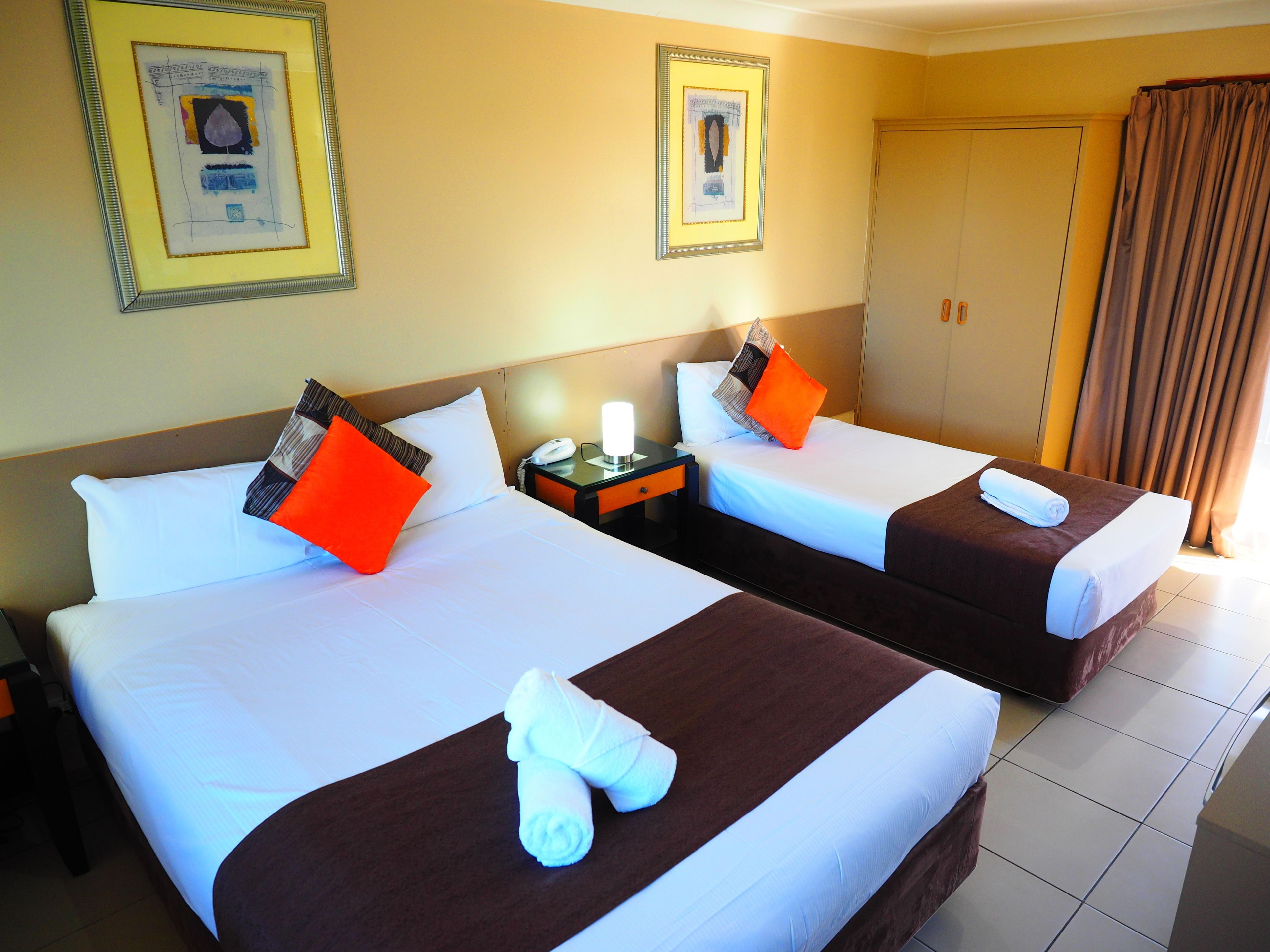 A hotel room with two beds, each with white linens and brown headboards. There are framed pictures on the walls, a nightstand with a lamp, and a door with curtains.
