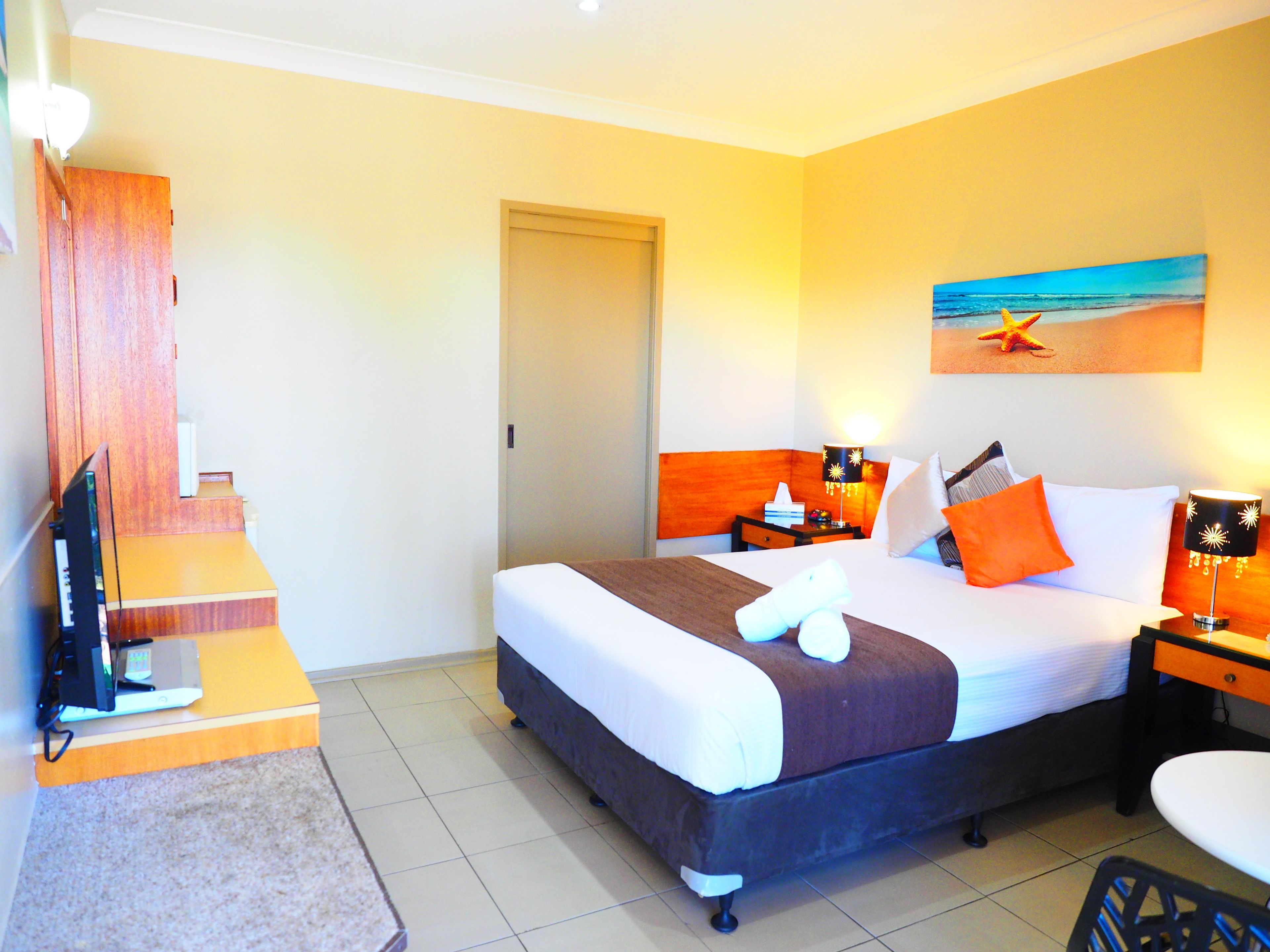 A hotel room with a bed, a desk, a chair, a TV, a painting of a beach, and a staircase.