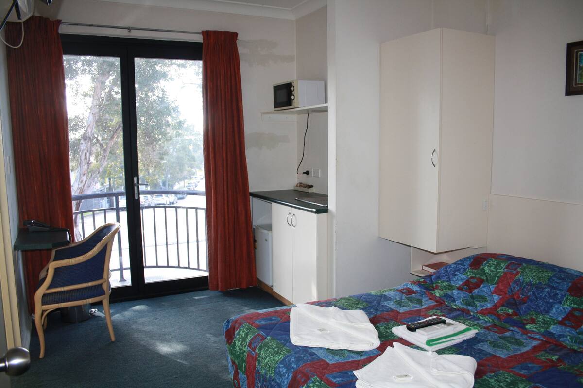 A hotel room with a bed, a desk, a chair, a wardrobe, and a view of the outside.