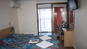 Quarto de hotel com uma cama, uma escrivaninha, uma cadeira, uma TV e uma porta de vidro deslizante para a varanda.
