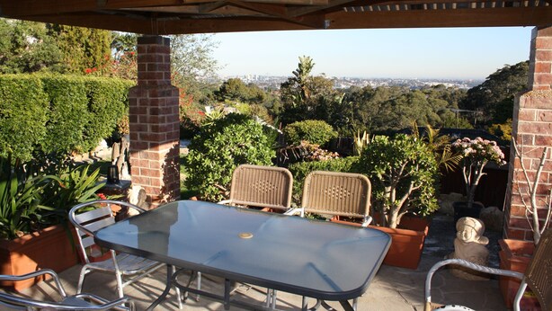 Um pátio externo com mesa e cadeiras, rodeado por plantas em vasos e vista para a paisagem urbana.