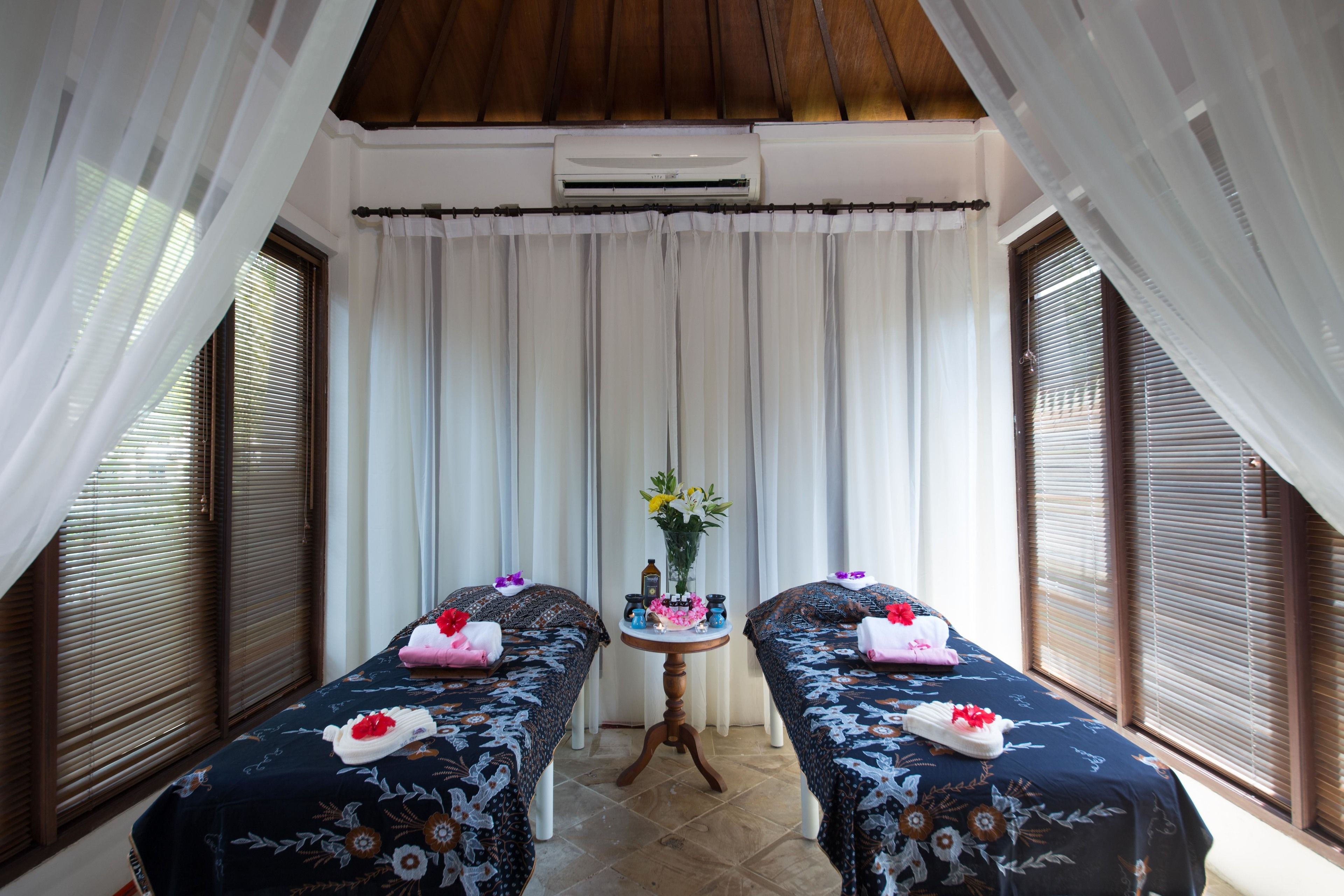 A room with two beds, a small table with flowers, and a window with blinds.