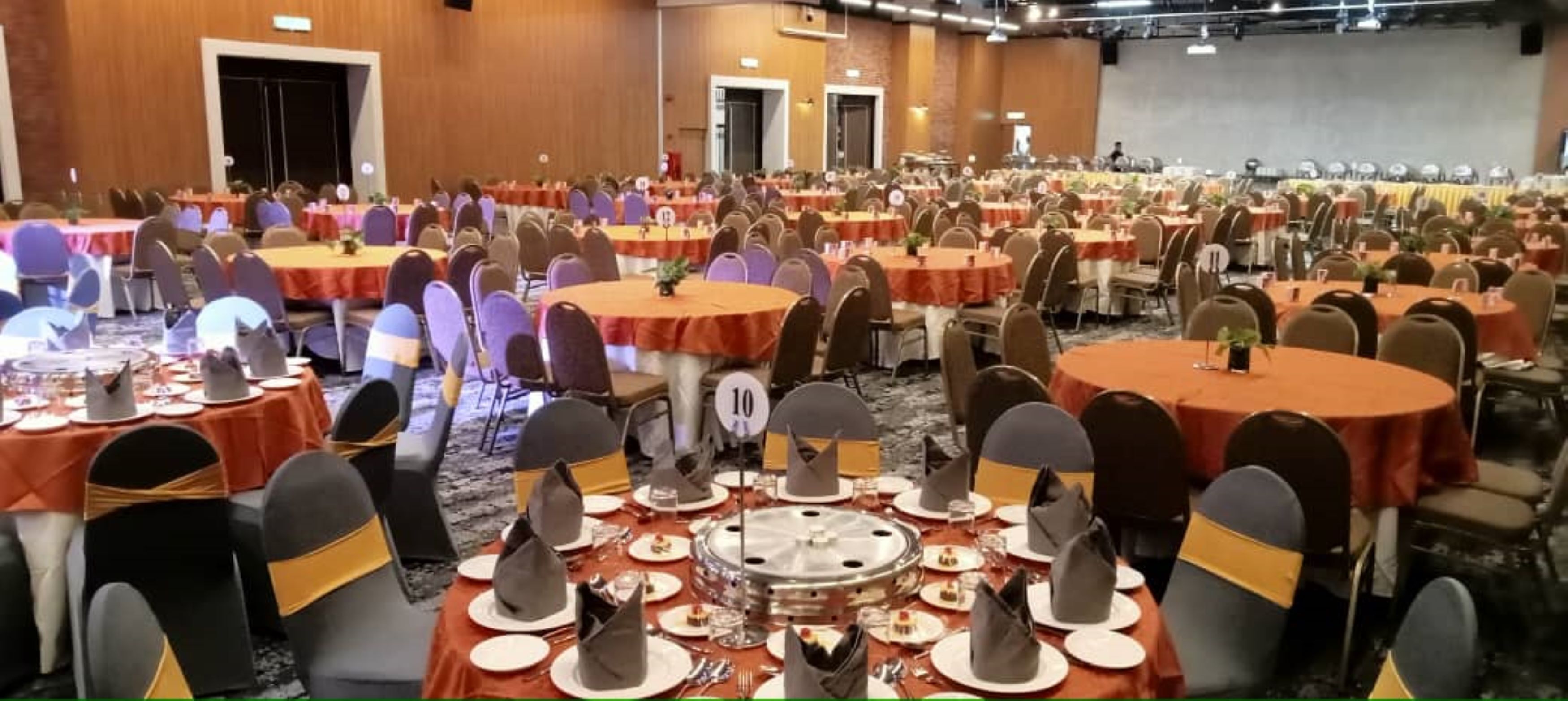 A large banquet hall with round tables covered in red tablecloths, each with a number sign, and chairs with black and gold sashes.
