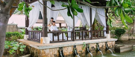 A gazebo with a stone base and white drapes, overlooking a water feature with multiple streams.