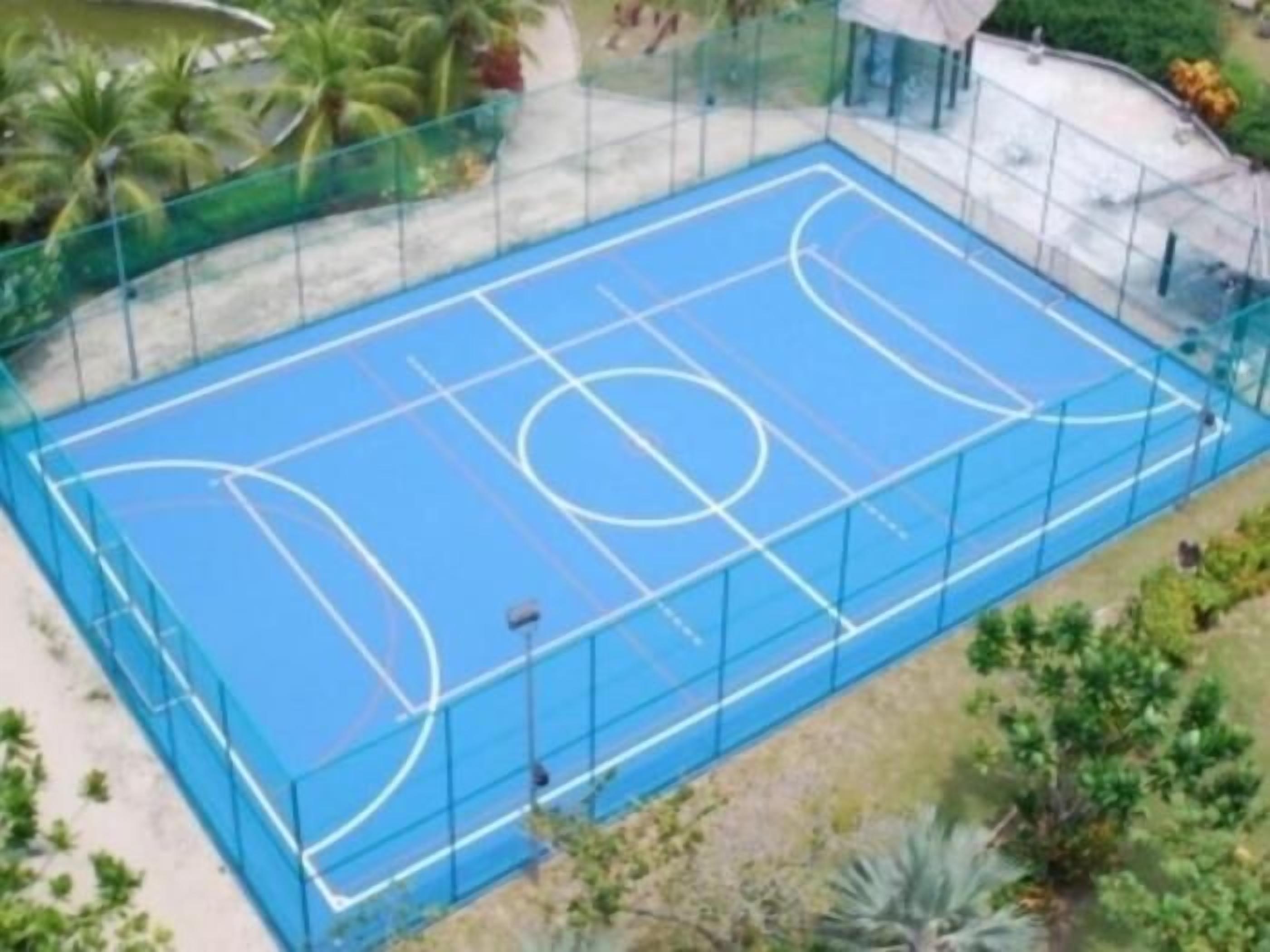 Una cancha de baloncesto al aire libre con superficie azul y líneas blancas, rodeada por una cerca verde y palmeras.