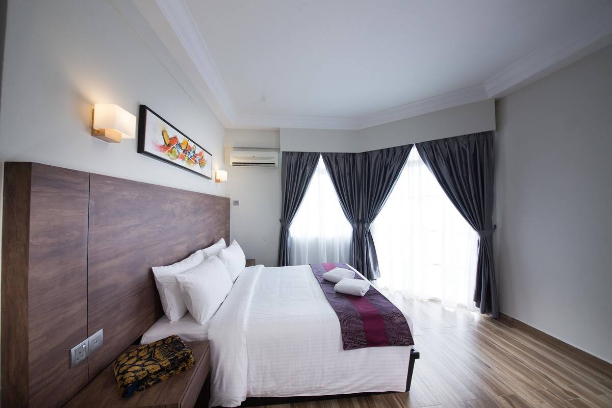 A modern hotel room with a large bed, a wooden headboard, a wall-mounted air conditioner, and a large window with curtains.