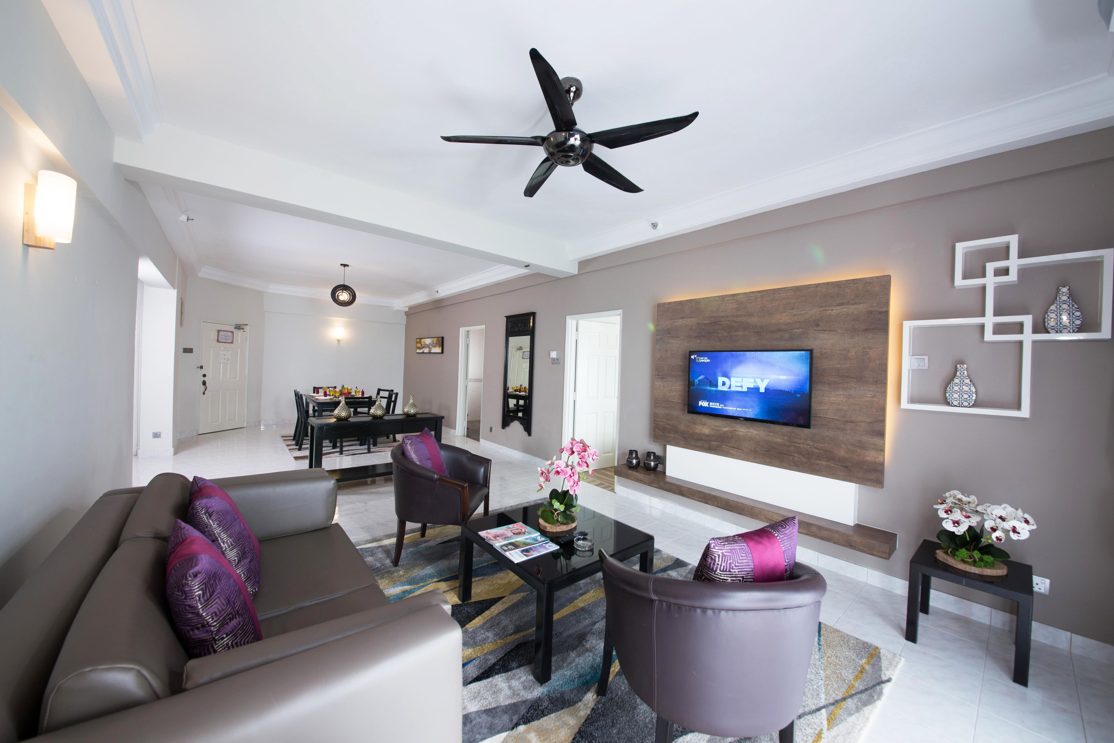 A modern living room with a ceiling fan, a flat-screen TV, a sofa, a coffee table, and a dining area in the background.