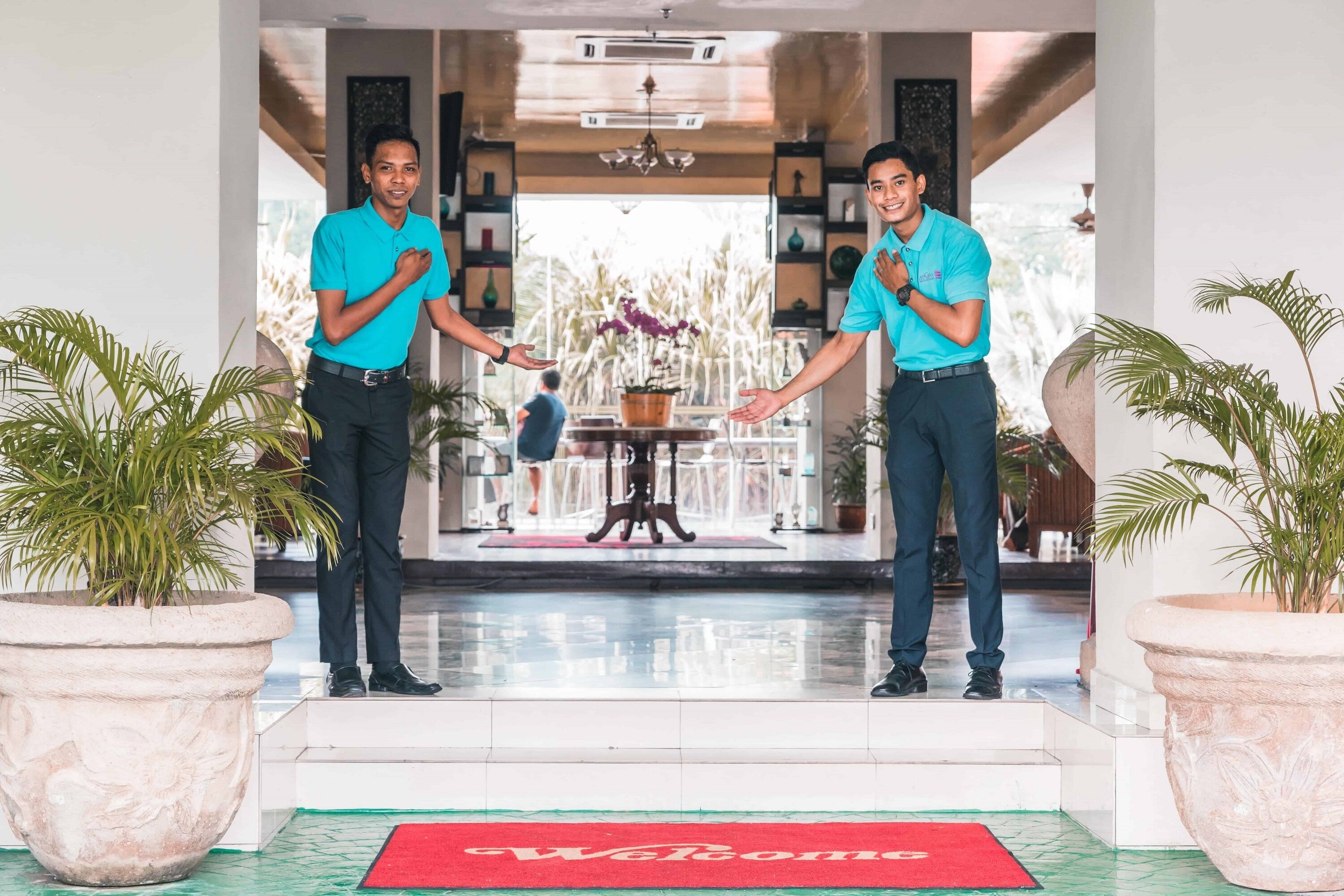 Two men in turquoise shirts shaking hands at a hotel entrance with a 'Welcome' mat, potted plants, and a view of the interior.