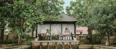 A serene garden with a small pavilion, a stone water feature, and lush greenery.