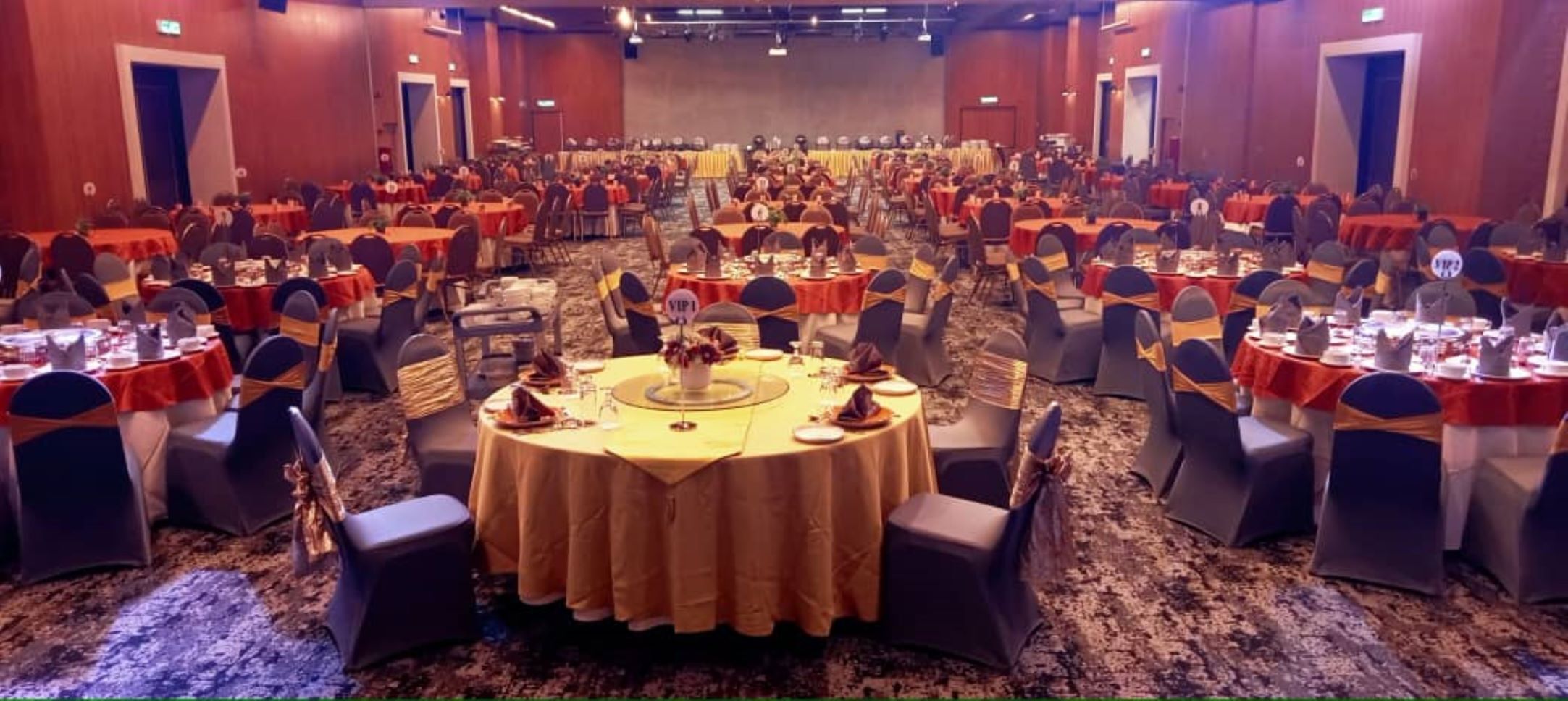 Uma sala de banquetes com mesas redondas, cadeiras e um palco ao fundo.