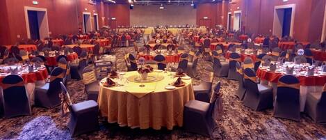 Uma sala de banquetes com mesas redondas, cadeiras e um palco ao fundo.