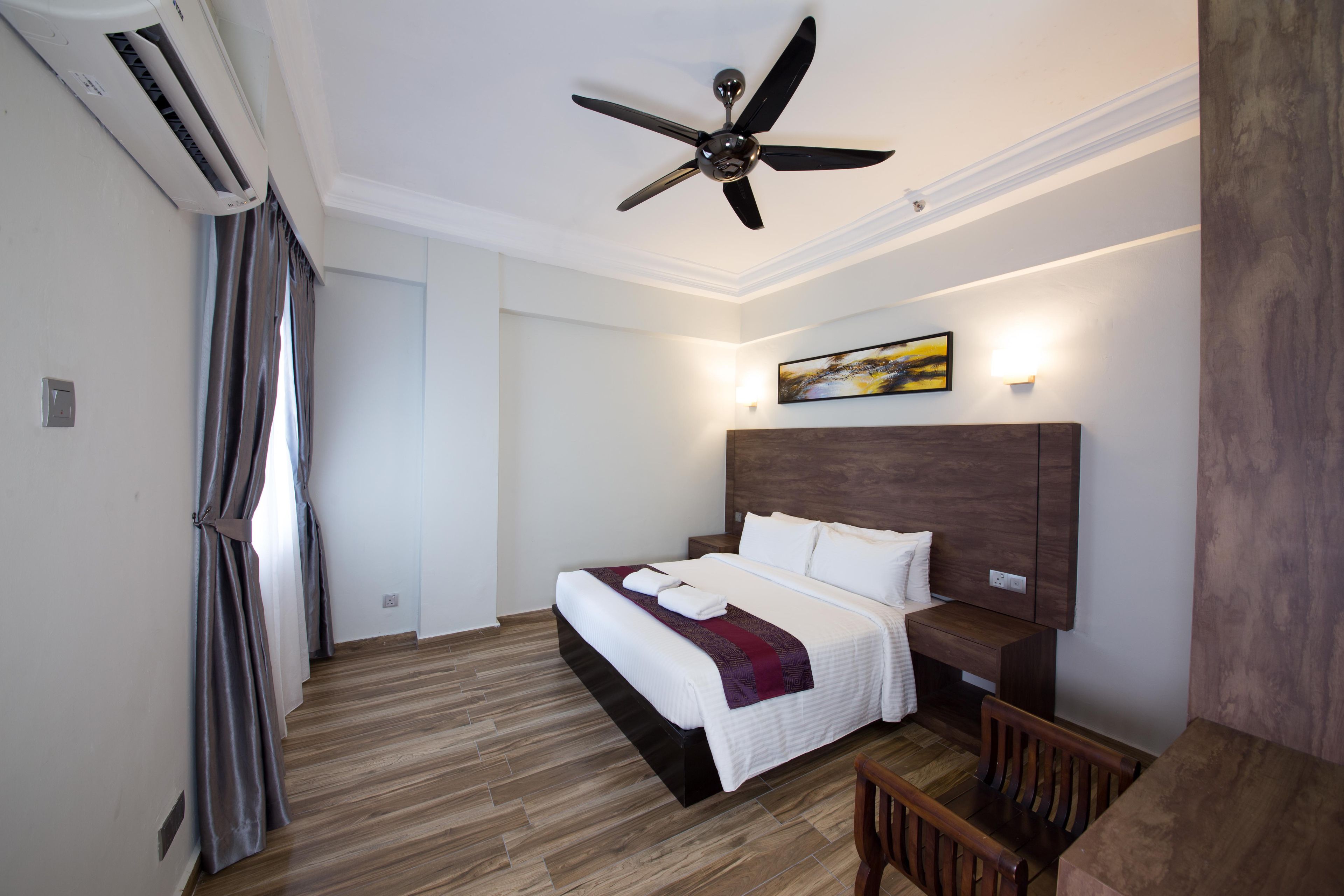 A hotel room with a bed, a wooden floor, a ceiling fan, an air conditioning unit, a nightstand, a bench, and a painting on the wall.