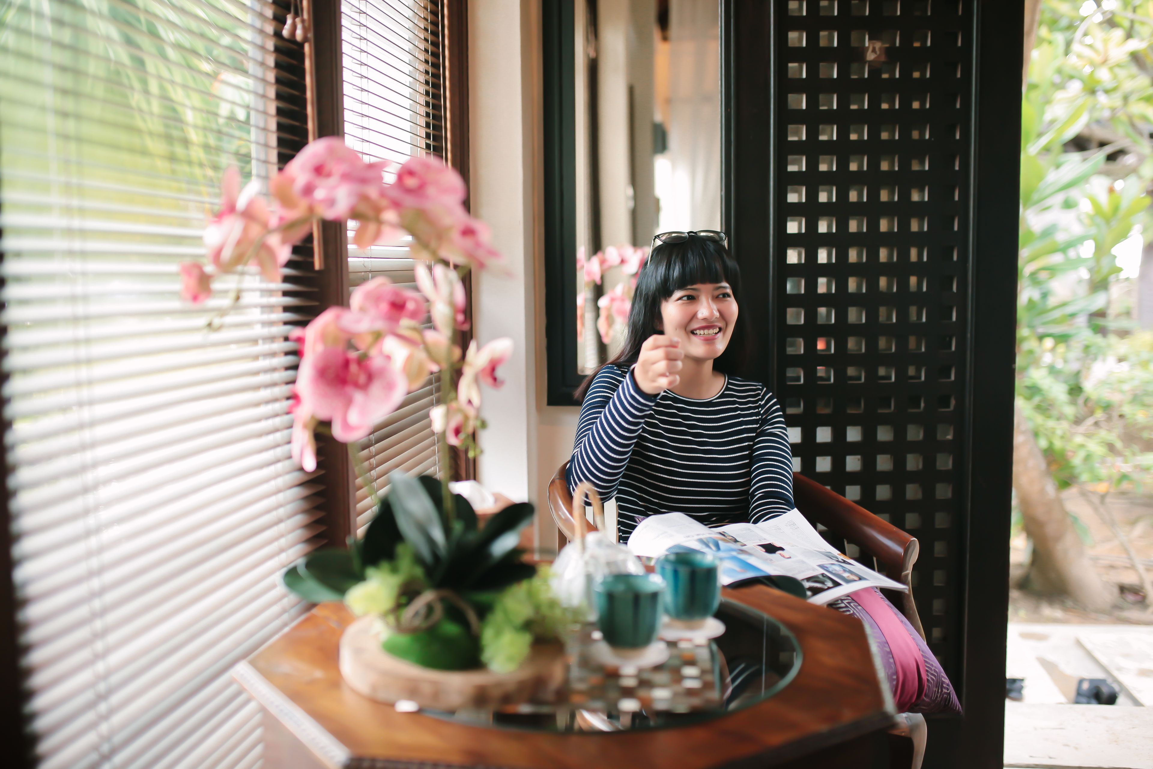 A person sitting at a wooden table with a book, a teapot, and a flower arrangement.