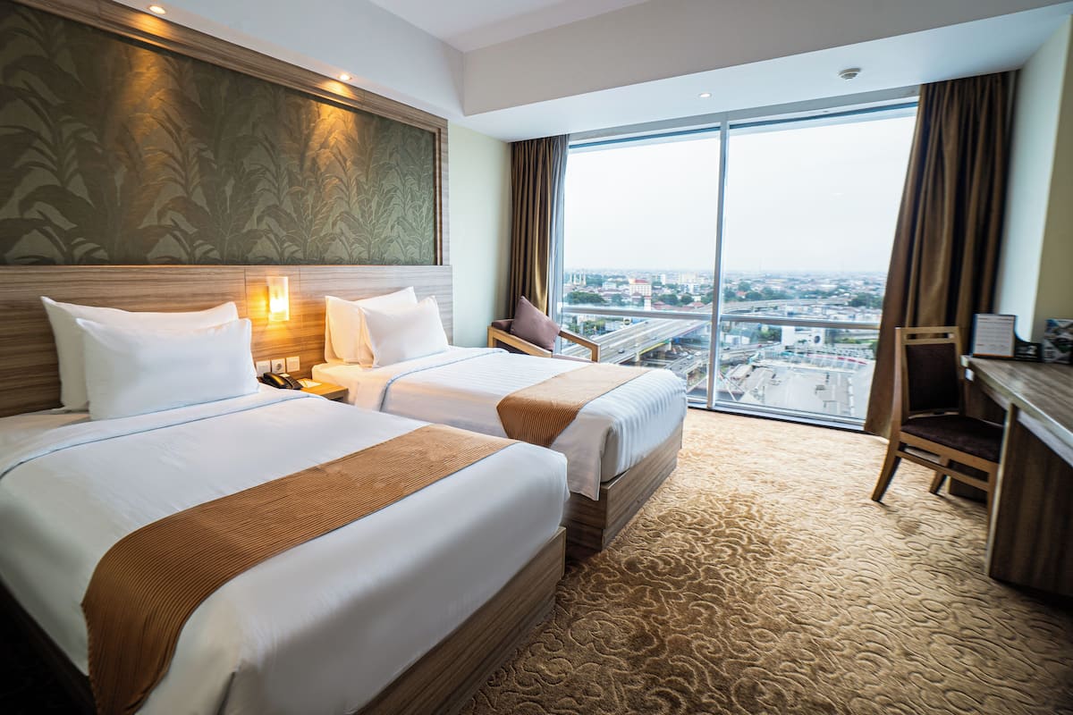 Ein Hotelzimmer mit zwei Betten, einem Schreibtisch und einem großen Fenster mit Blick auf die Stadt.