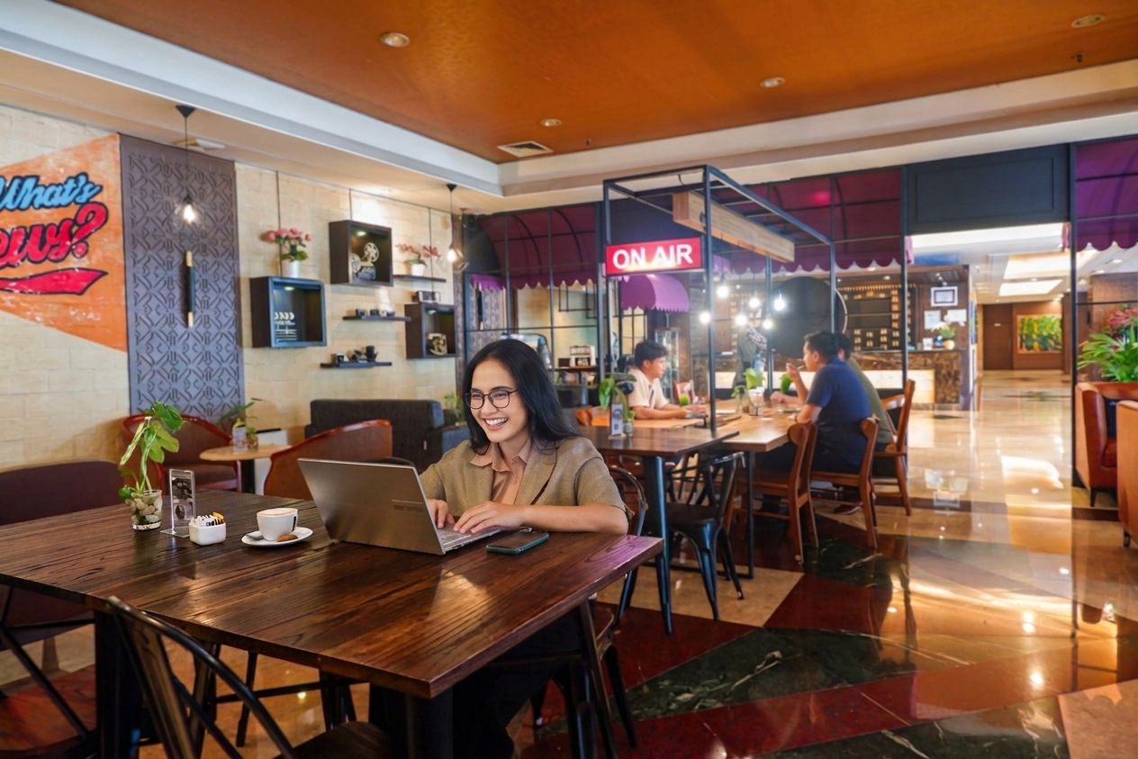 A modern indoor dining area with wooden tables and chairs, a visible 'ON AIR' sign, and a person using a laptop.