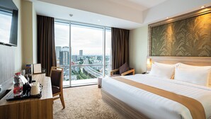 A hotel room with a large bed, a desk with a coffee maker, a chair, and a city view through large windows. - Horison Ultima Bekasi (Bekasi)
