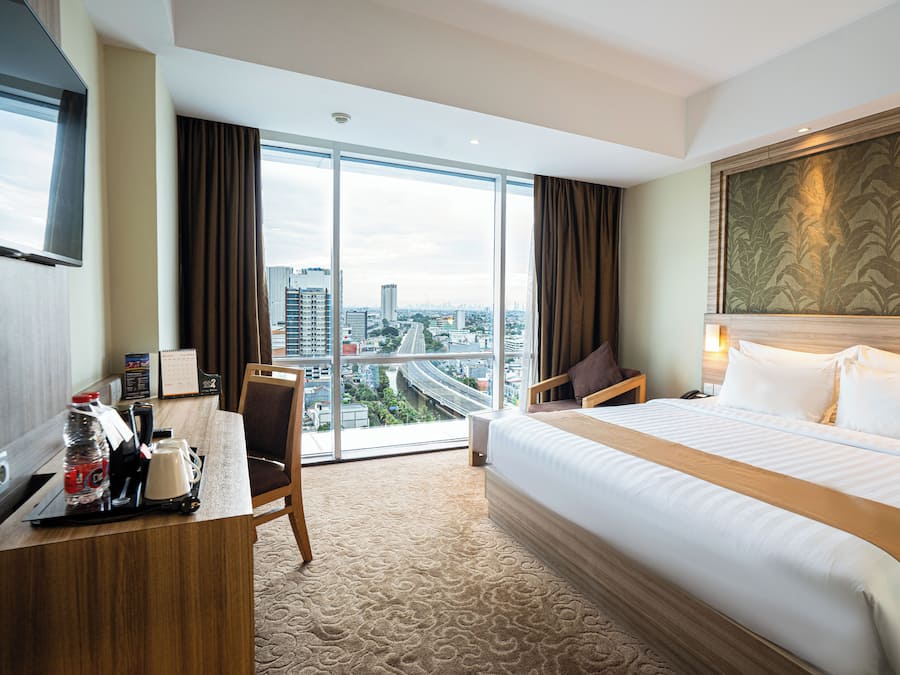 Habitación de hotel con una cama grande, un escritorio con cafetera, una silla y vistas a la ciudad a través de amplias ventanas.