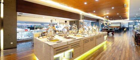 A buffet area with a variety of food options, a counter with plates, and a view of the cityscape through large windows.