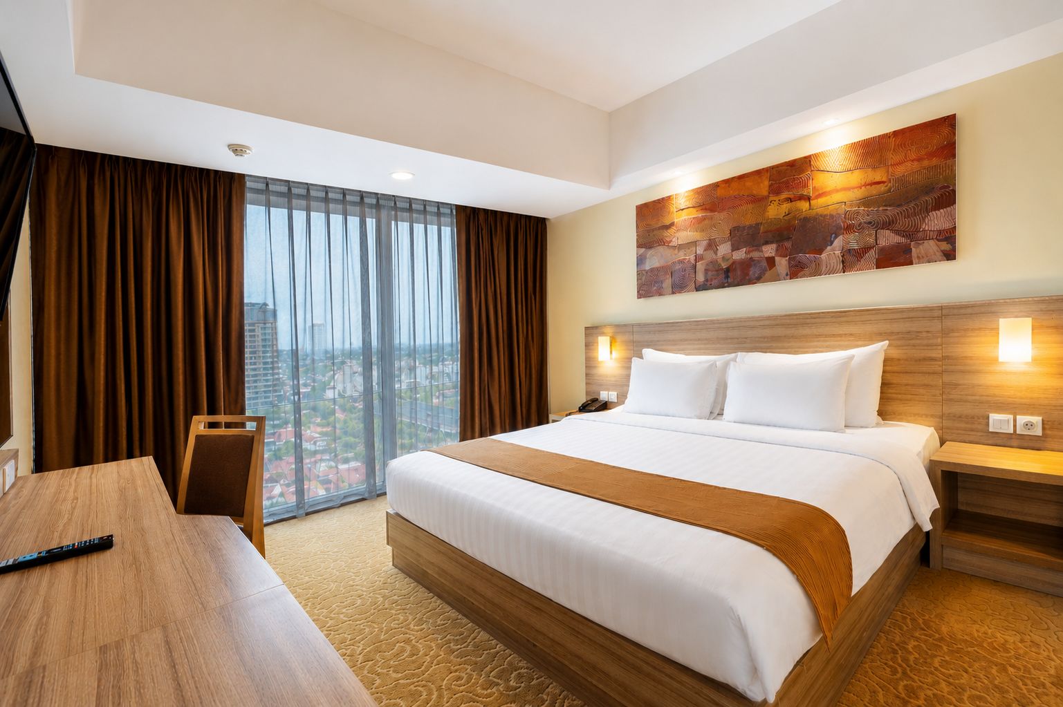 A hotel room with a large bed, a desk, a chair, a view of the city, and a painting on the wall.