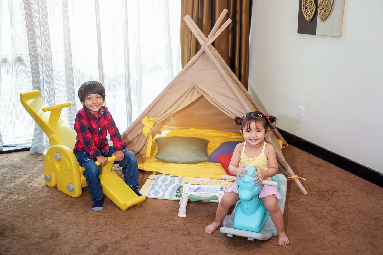 Dos niños jugando en una habitación con una carpa de juegos, un tobogán amarillo y un caballo de juguete.