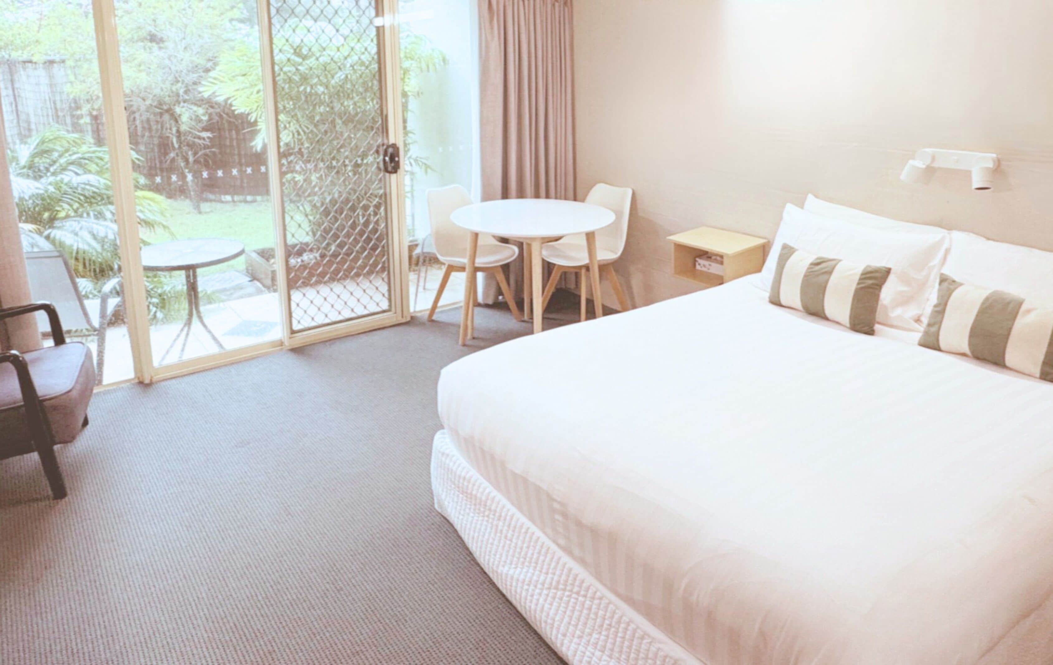 A hotel room with a large bed, a small table, two chairs, and a sliding glass door leading to an outdoor area with a view of greenery.