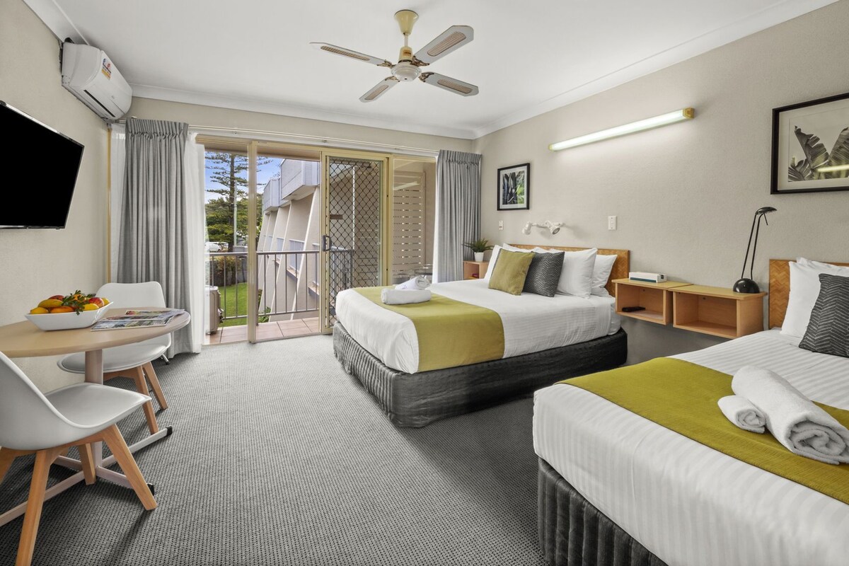 Habitación de hotel con cama, escritorio, silla, televisión, ventilador de techo y balcón con cortinas.