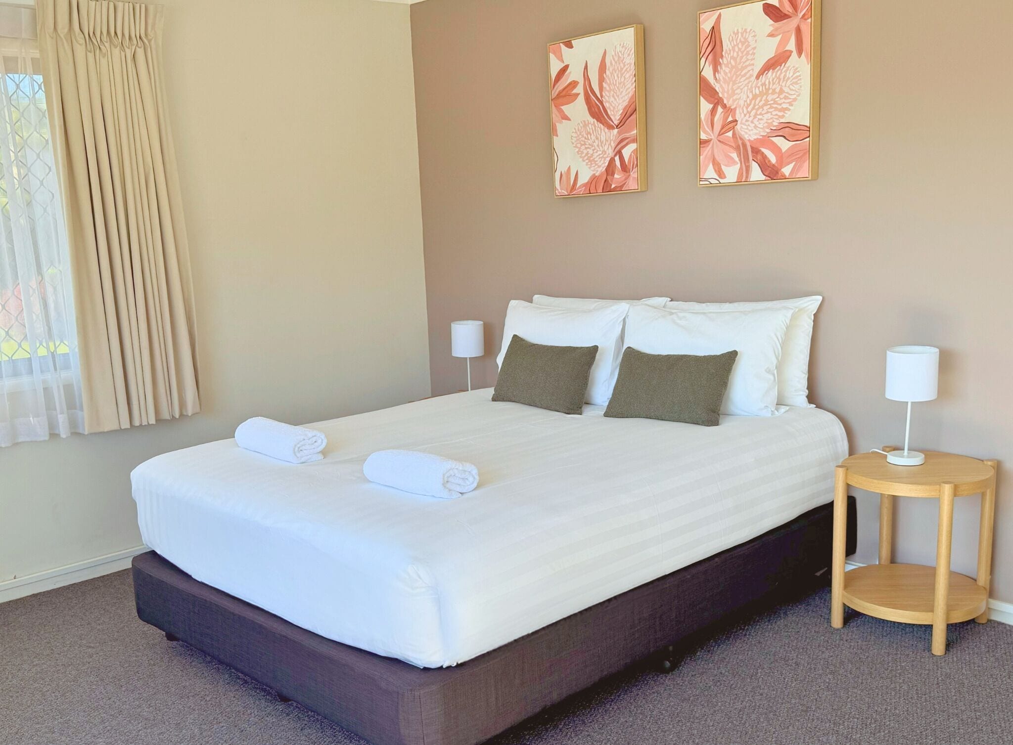 Una cama bien hecha con sábanas blancas y dos almohadas, una mesita de noche con una lámpara y dos cuadros florales en la pared.