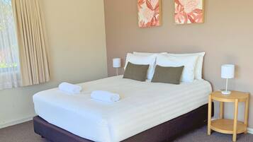 Una cama bien hecha con sábanas blancas y dos almohadas, una mesita de noche con una lámpara y dos cuadros florales en la pared.