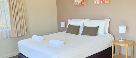 Una cama bien hecha con sábanas blancas y dos almohadas, una mesita de noche con una lámpara y dos cuadros florales en la pared.