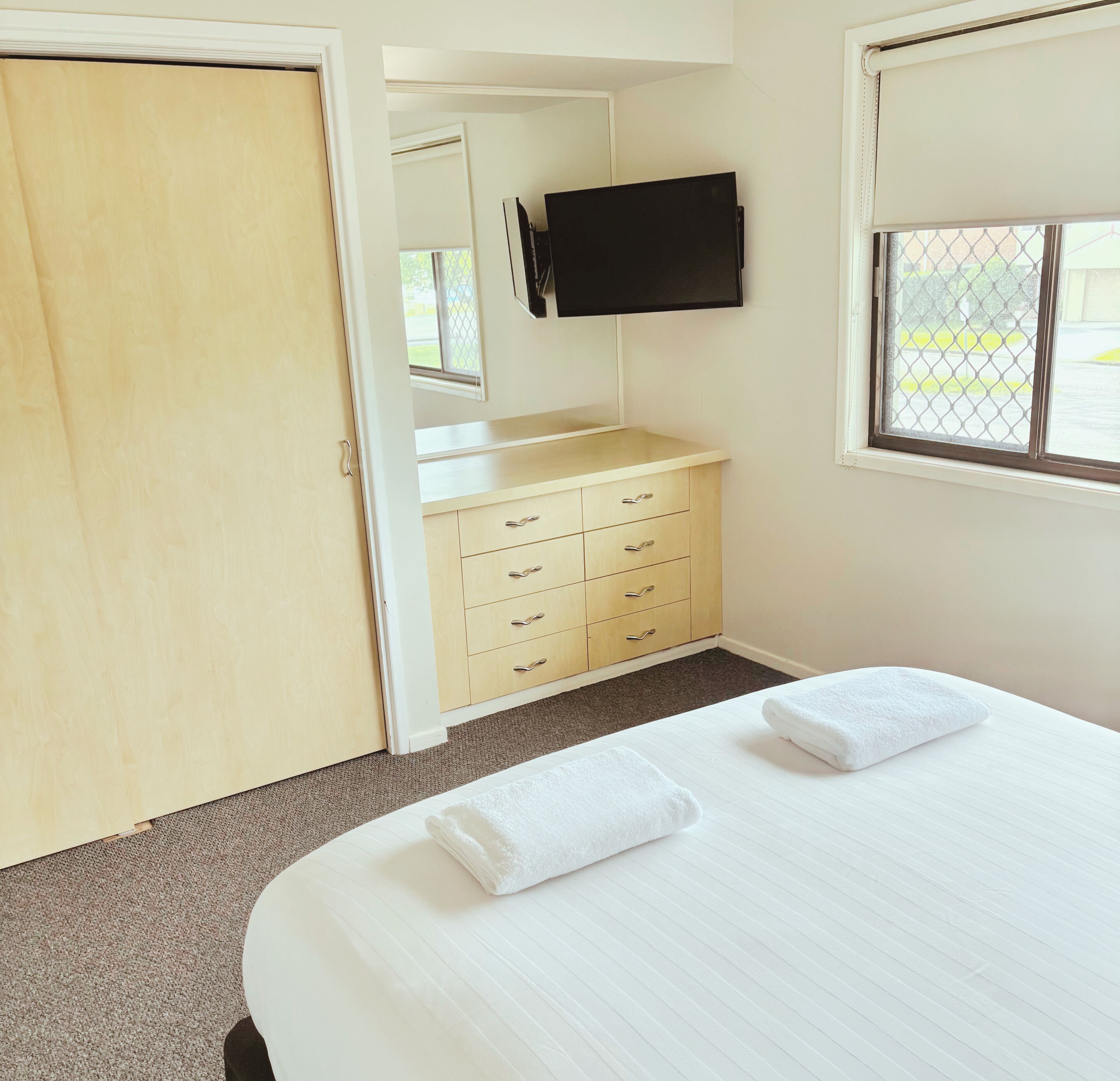 A hotel room with a bed, a wardrobe, a TV mounted on the wall, and a window with a view of the outside.