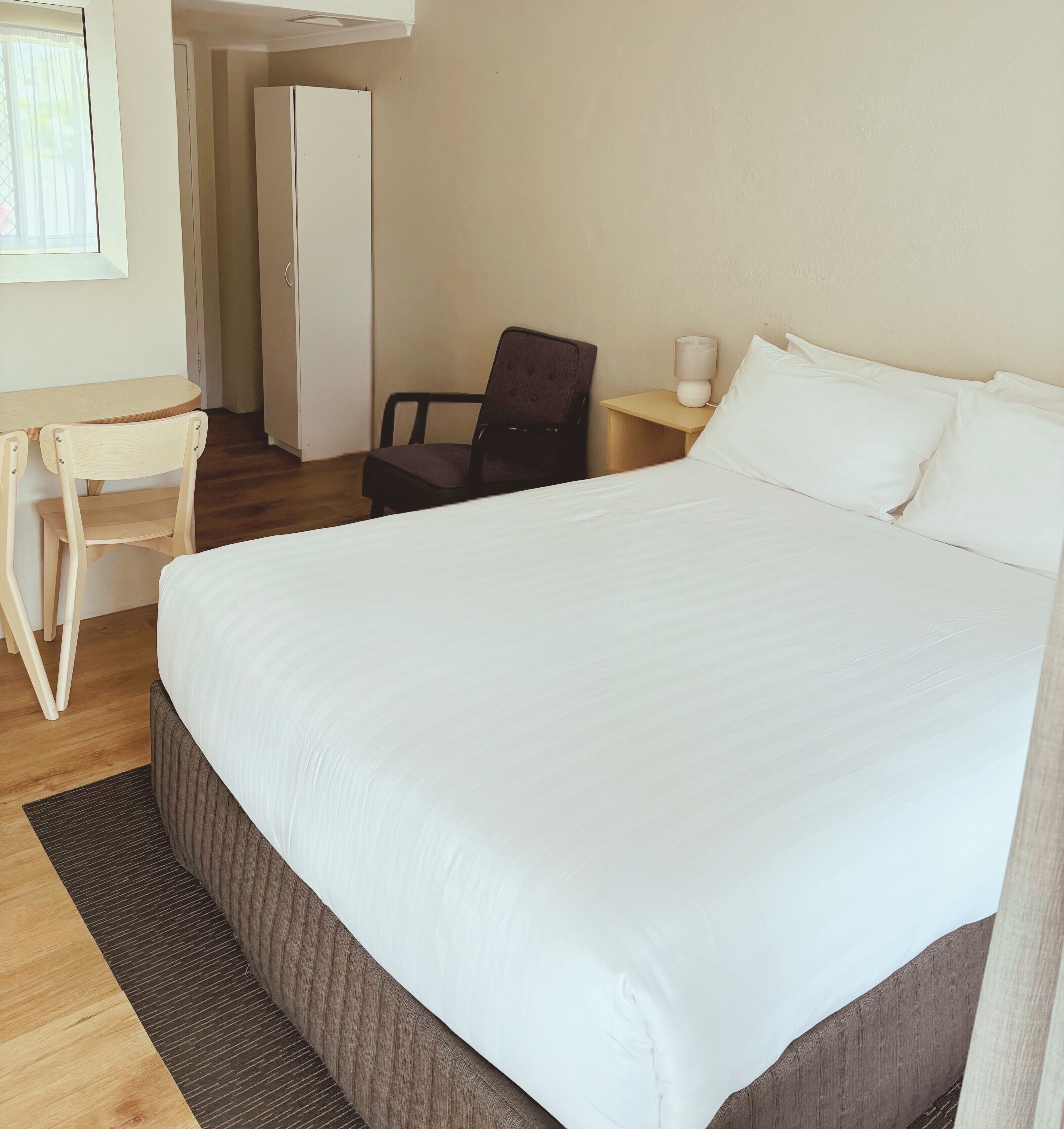 A neatly arranged hotel room with a large bed, a desk, a chair, and a bedside table with a lamp.